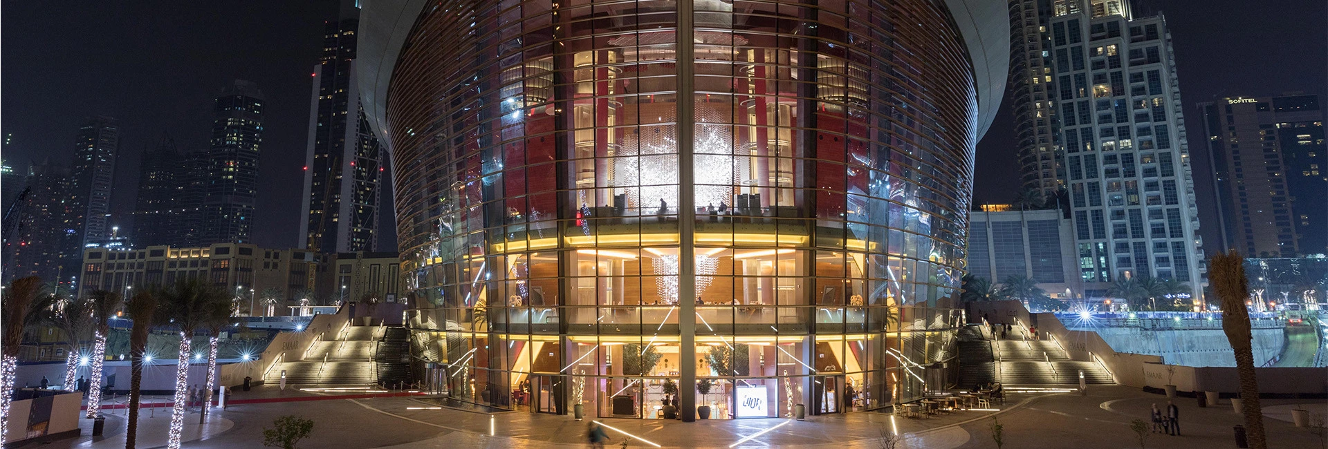 Illuminated Dubai Opera building at night, surrounded by skyscrapers.