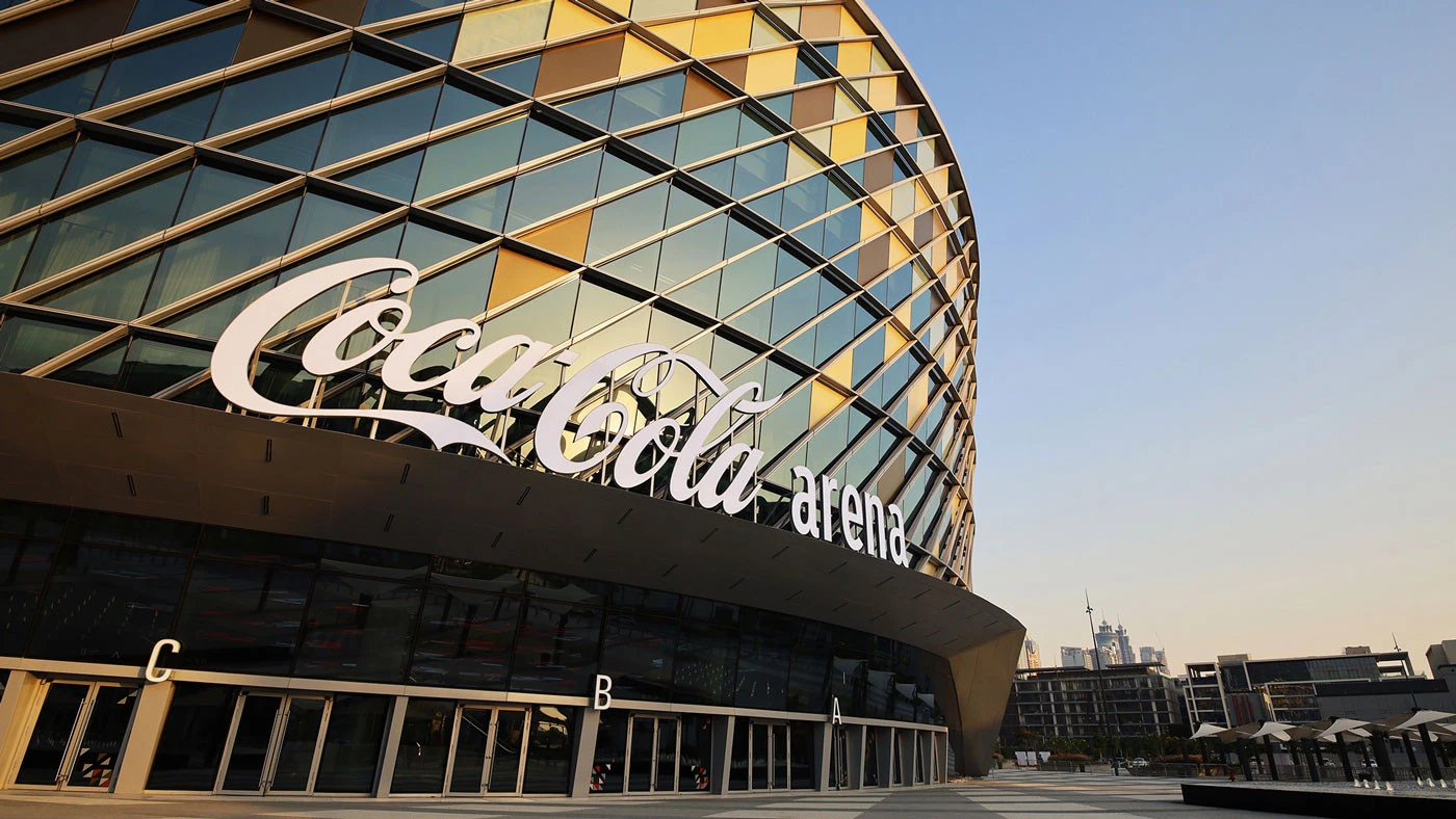 Coca-Cola Arena exterior with glass facade, Dubai skyline in background.