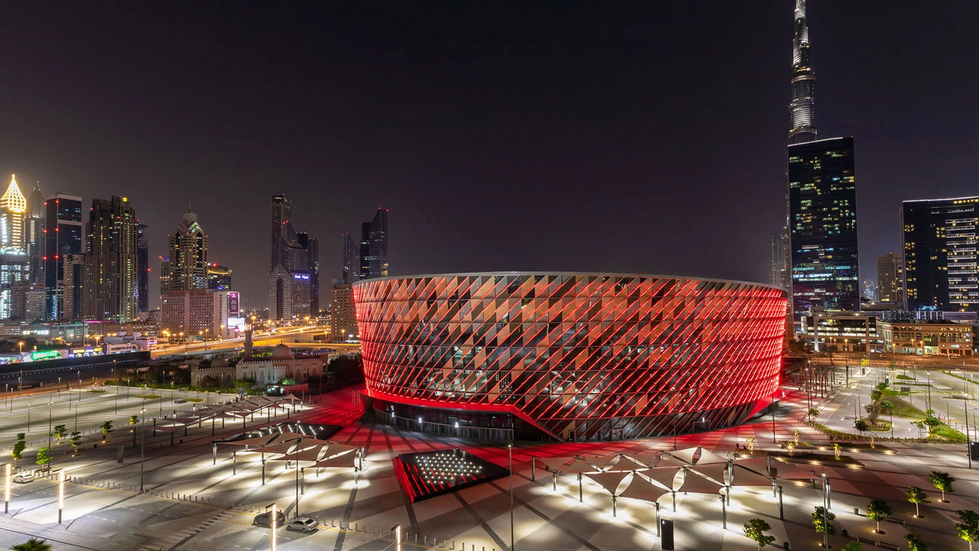 Coca-Cola Arena illuminated at night, Dubai skyline in background.