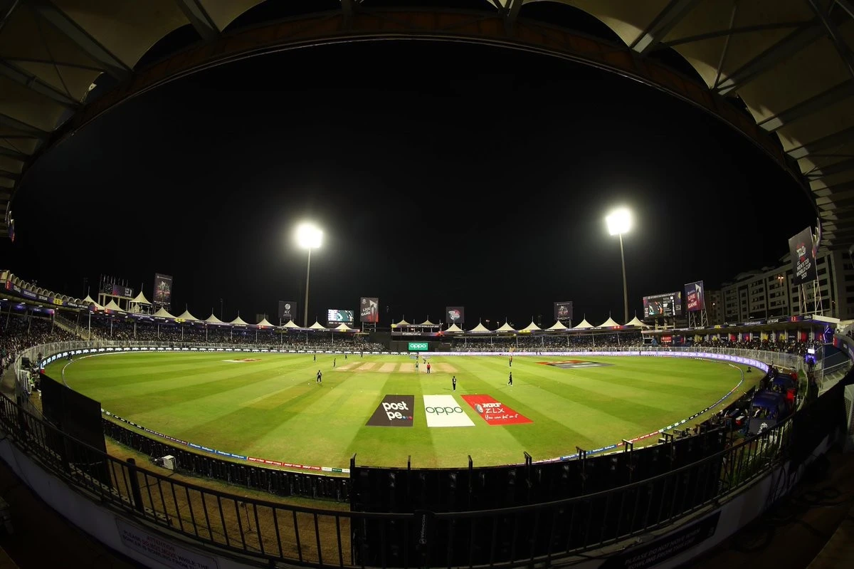 Sharjah Cricket Stadium illuminated during nighttime event, Sharjah cityscape visible.