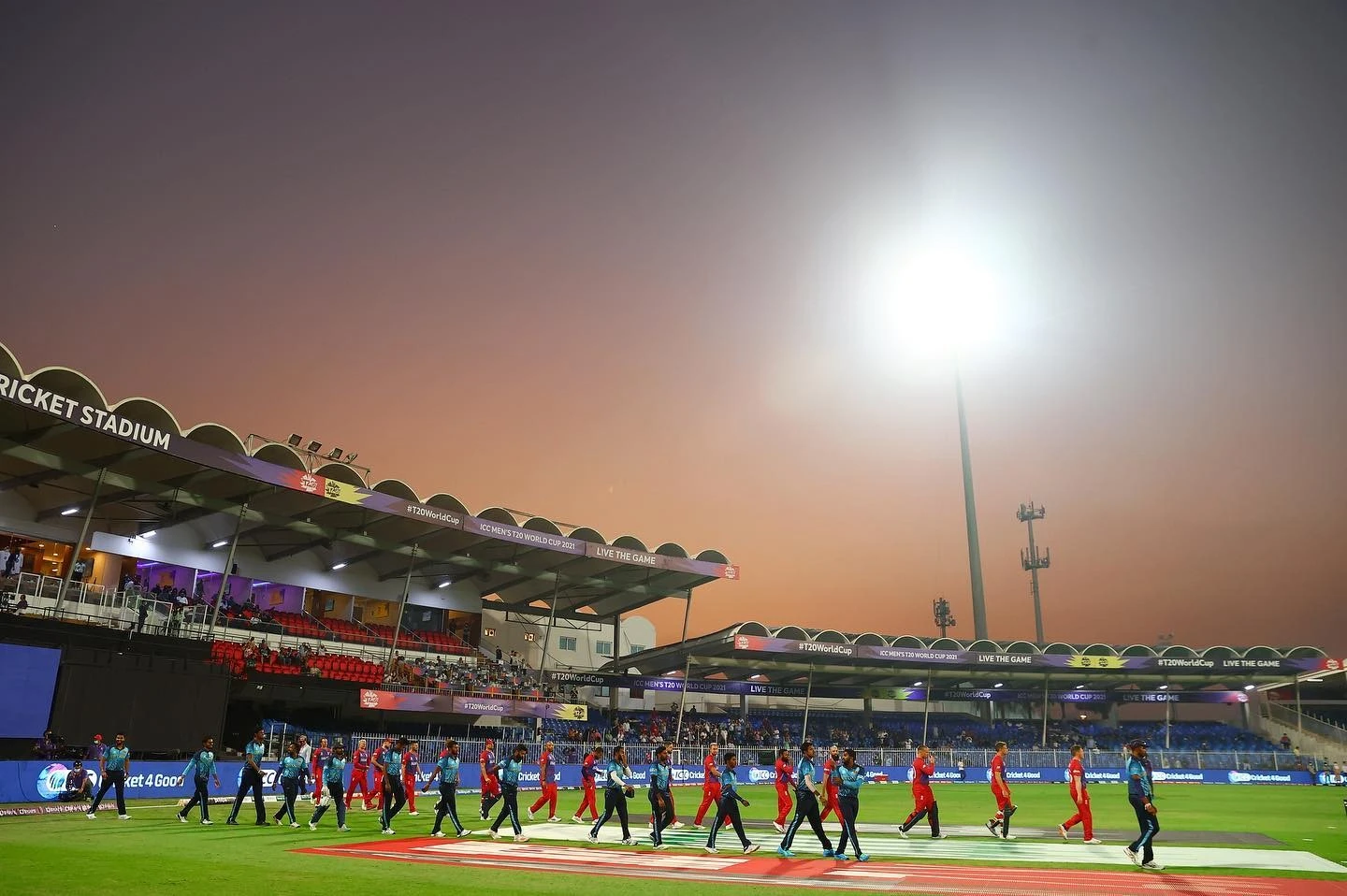 Players walking on Sharjah Cricket Stadium field at sunset.